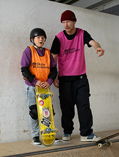 Skate Academy leerling met hesje en een gouden skateboard links, met begeleider Bram naast hem die uitleg geeft.